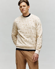 A man with light brown hair wears the Psycho Bunny MENS REED FLOCK CREW pullover (MT0500041) with a subtle hand gesture pattern and tan cargo pants, standing against a plain light gray background, one hand in his pocket.