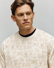 A man with short brown hair wears the Psycho Bunny MENS REED FLOCK CREW (MT0500041), a premium cotton-blend pullover featuring cartoon bunny faces, skulls, and "PB," as he looks slightly to the side against a plain light background.
