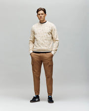 A man faces forward with hands in pockets, wearing Psycho Bunny’s MENS REED FLOCK CREW - MT0500041 beige pullover sweater, brown cargo pants, and black Adidas sneakers against a plain light gray background.