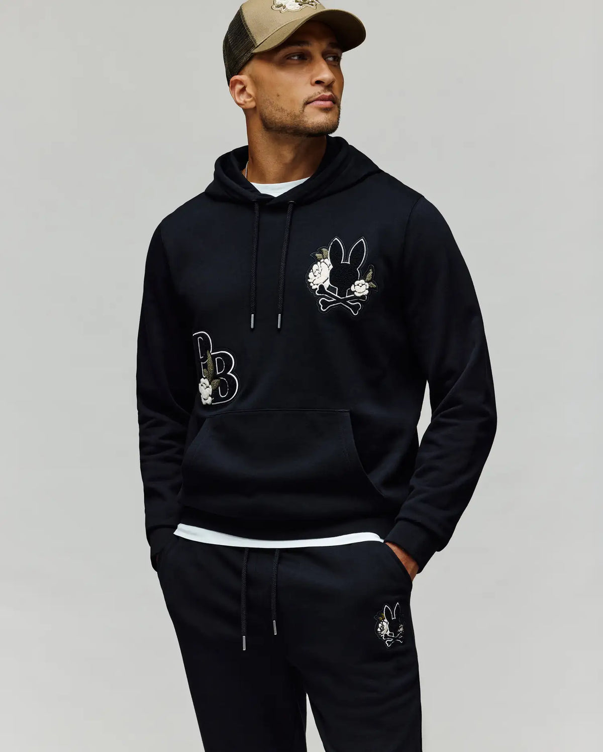A man stands with hands in his pockets, wearing the Psycho Bunny MENS AUGUSTE HOODIE - MT0500043 and matching sweatpants with cartoon bunny and chenille floral details, plus a beige and black baseball cap, looking off to the side.