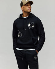 A man stands with hands in his pockets, wearing the Psycho Bunny MENS AUGUSTE HOODIE - MT0500043 and matching sweatpants with cartoon bunny and chenille floral details, plus a beige and black baseball cap, looking off to the side.