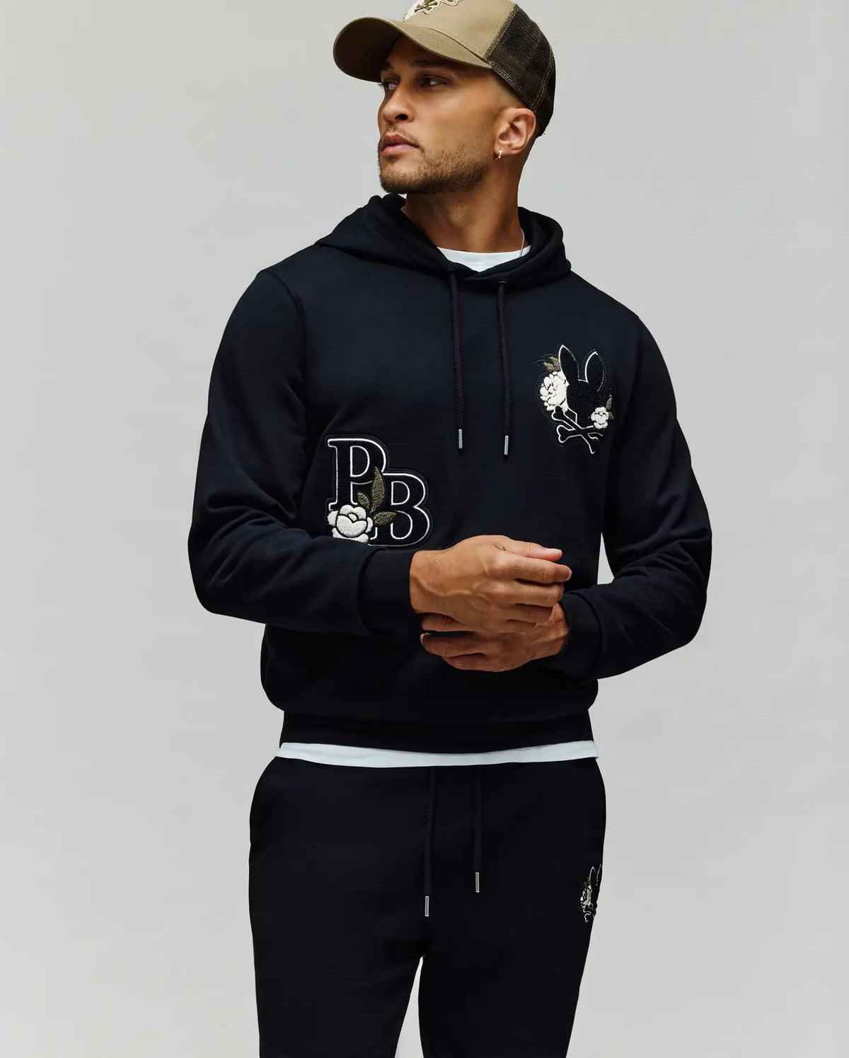 A man in the Psycho Bunny MENS AUGUSTE HOODIE (MT0500043) and matching joggers—both with embroidered bunny and "PB" logos—wears a green-brown cap, looking to the side against a plain background.