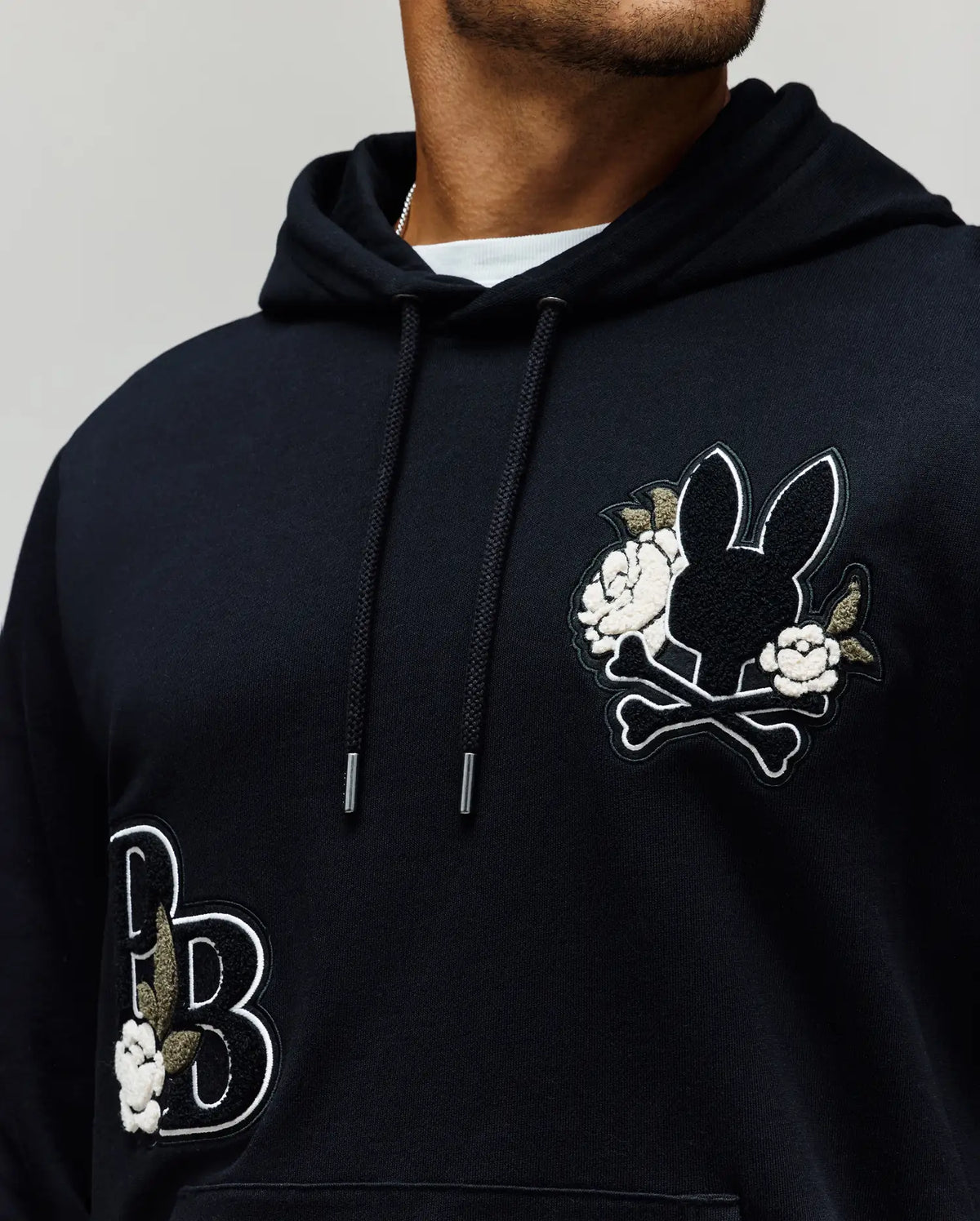 A person wears the Psycho Bunny MENS AUGUSTE HOODIE (MT0500043), a regular fit premium cotton hoodie with embroidered patches—a rabbit head with crossbones and flowers on the chest, plus chenille floral and "PB" accents on the lower left.