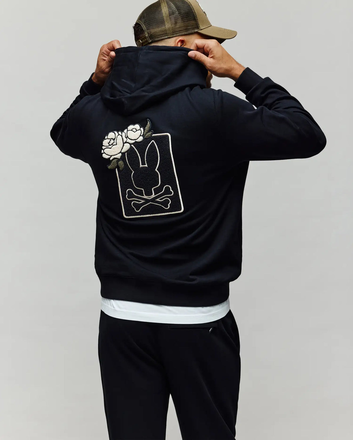 A person in a Psycho Bunny MENS AUGUSTE HOODIE (MT0500043) and cap faces away, showing a back graphic with a bunny silhouette, crossbones, two white roses, and chenille floral details.