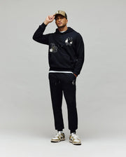 A man stands against a plain background wearing the Psycho Bunny MENS AUGUSTE HOODIE (MT0500043), matching joggers with cartoon graphics, a tan baseball cap, and white sneakers with beige accents, holding his cap's brim with one hand.