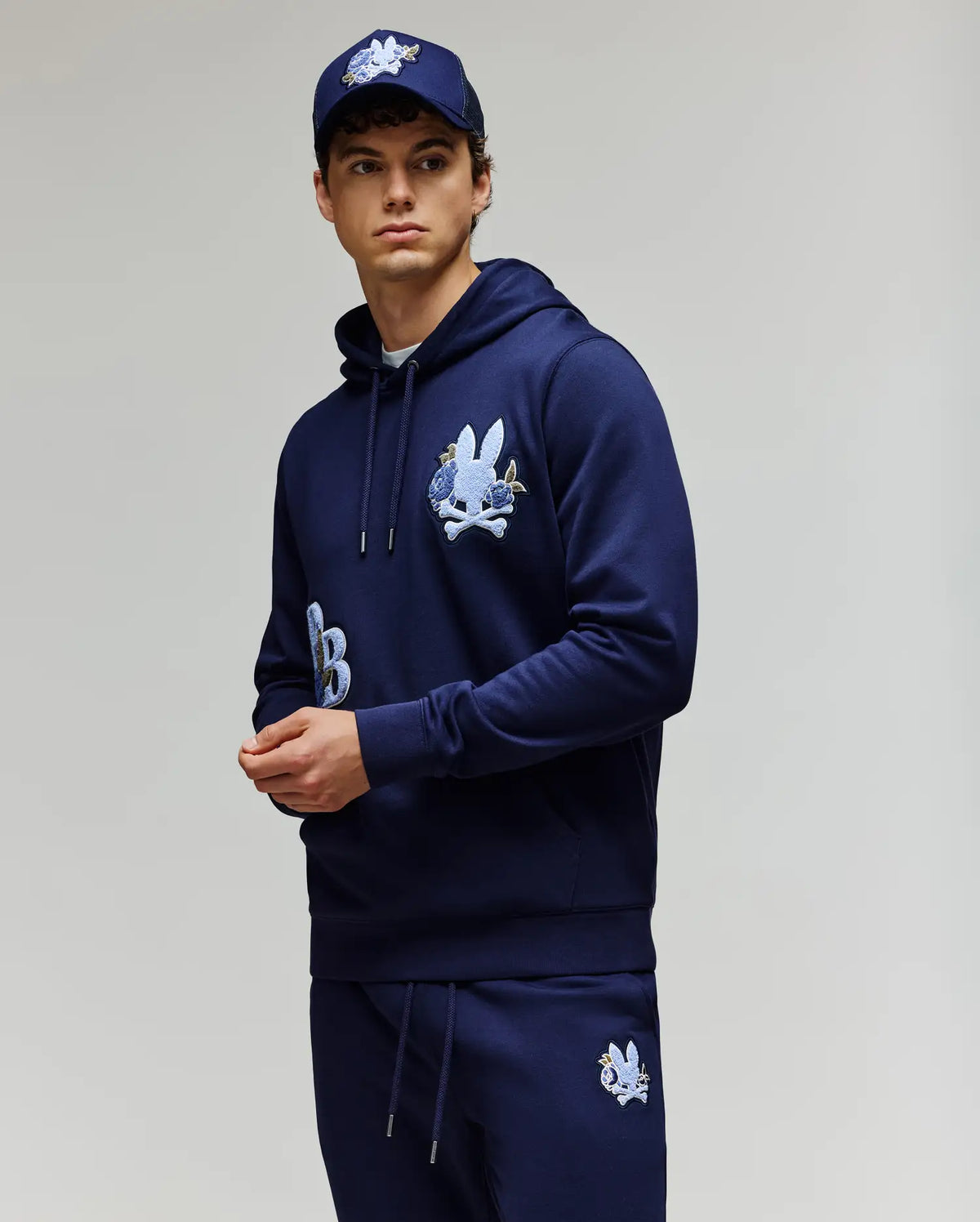 A young man wears the Psycho Bunny MENS AUGUSTE HOODIE (MT0500043) in regular fit navy blue premium cotton with chenille floral detailing, matching sweatpants, and a navy baseball cap against a plain light background.