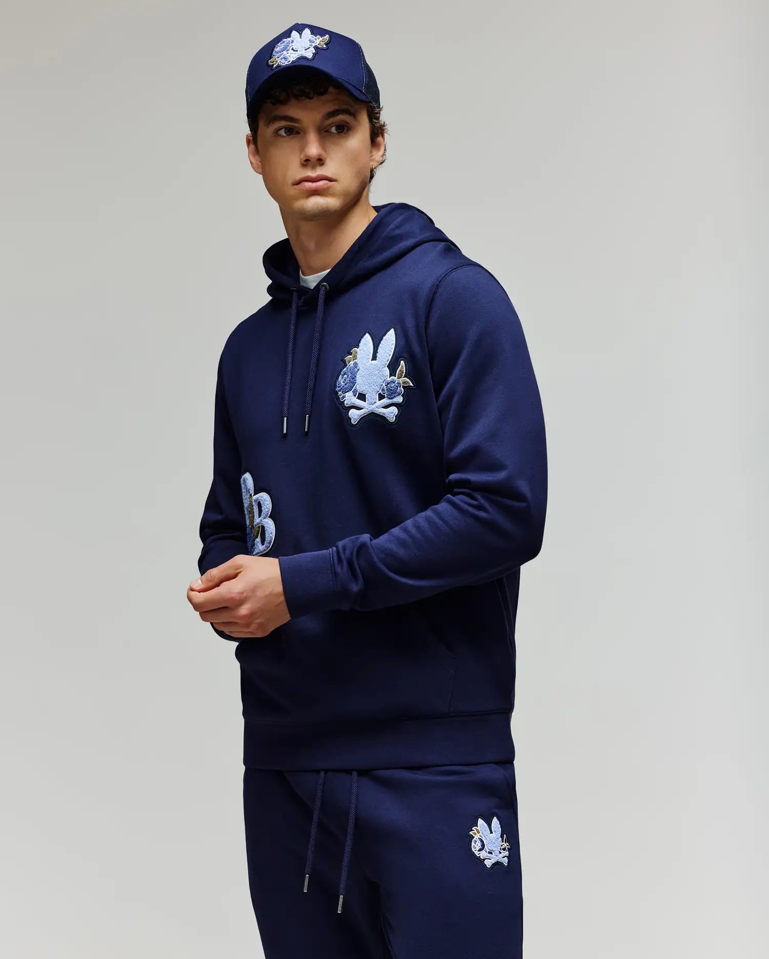 A young man wears the Psycho Bunny MENS AUGUSTE HOODIE (MT0500043) in regular fit navy blue premium cotton with chenille floral detailing, matching sweatpants, and a navy baseball cap against a plain light background.
