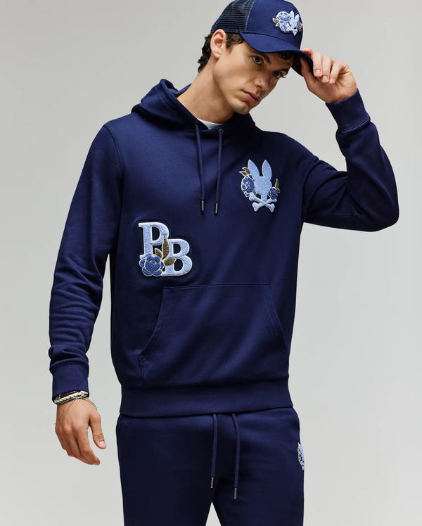A man wears the Psycho Bunny MENS AUGUSTE HOODIE - MT0500043 in navy, matching joggers with chenille floral details, and a navy cap, tilting it while looking at the camera against a plain background.
