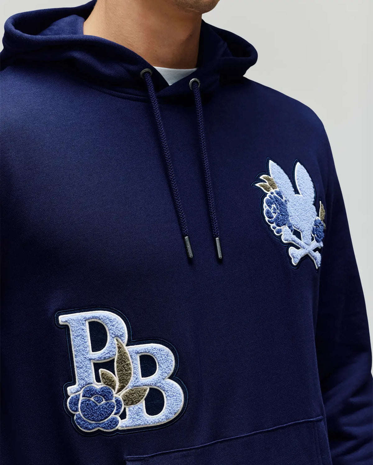 Wearing the Psycho Bunny MENS AUGUSTE HOODIE - MT0500043 in navy blue, featuring premium cotton, chenille floral accents, large "PB" chest lettering, and a patch with a stylized rabbit and flowers. Only the lower face and upper body are shown.