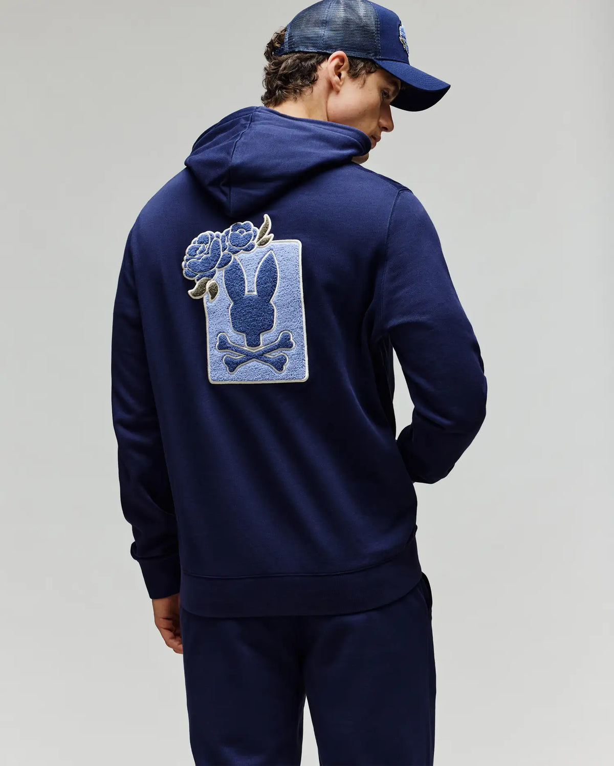 A person in Psycho Bunny’s MENS AUGUSTE HOODIE (MT0500043) and matching pants faces away, showing a bunny skull patch with blue chenille flowers. The outfit is finished with a dark blue cap.