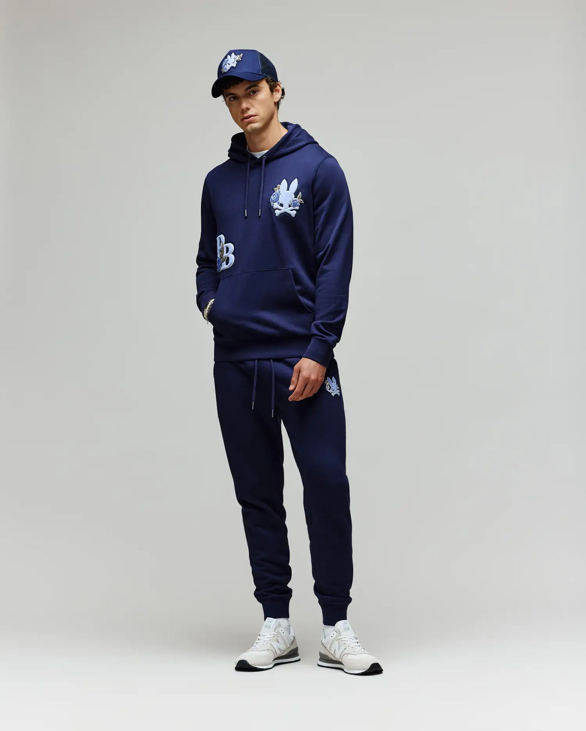 A person stands against a plain background wearing the Psycho Bunny MENS AUGUSTE HOODIE (MT0500043) with chenille floral detailing, matching joggers, a navy baseball cap, and white sneakers. Their hands are in their pockets and they look relaxed.