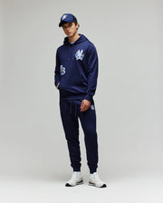 A person stands against a plain background wearing the Psycho Bunny MENS AUGUSTE HOODIE (MT0500043) with chenille floral detailing, matching joggers, a navy baseball cap, and white sneakers. Their hands are in their pockets and they look relaxed.