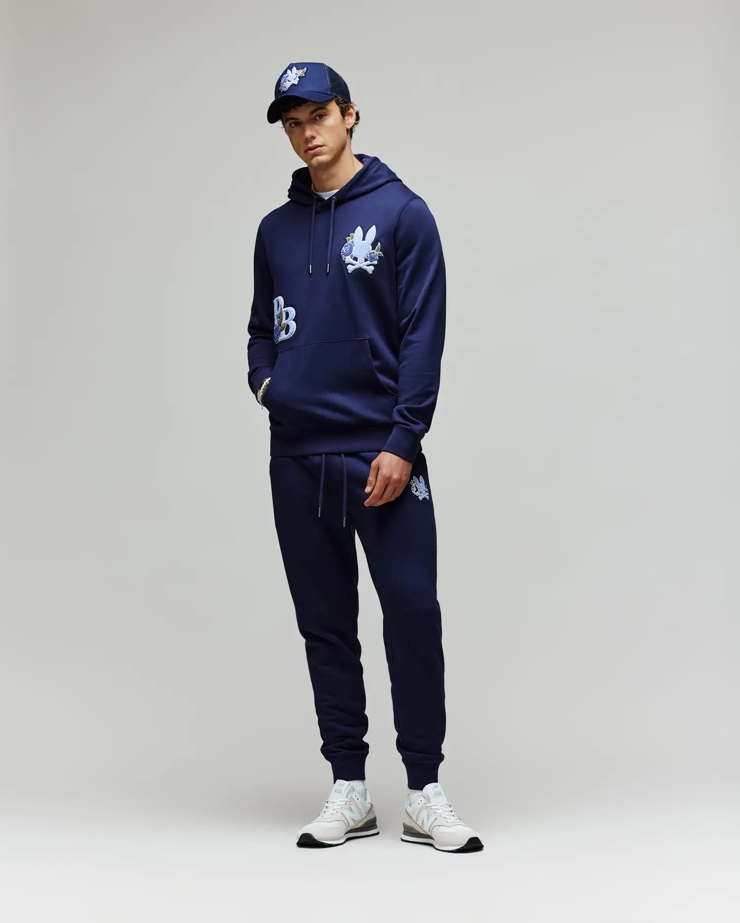 A person stands against a plain background wearing the Psycho Bunny MENS AUGUSTE HOODIE (MT0500043) with chenille floral detailing, matching joggers, a navy baseball cap, and white sneakers. Their hands are in their pockets and they look relaxed.