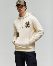 A young man stands against a plain background, wearing Psycho Bunny's MENS AUGUSTE HOODIE (MT0500043) with a bunny patch, a matching beige cap, and dark pants, hands in the front pocket as he looks at the camera.
