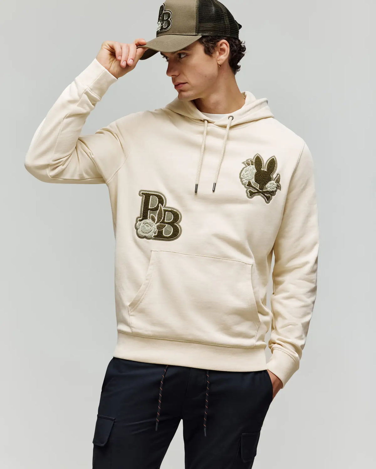A man wearing the Psycho Bunny MENS AUGUSTE HOODIE (MT0500043) in light beige, featuring "PB" chenille floral detail, pairs it with black pants and a black-and-khaki baseball cap, standing with one hand holding the cap's brim.