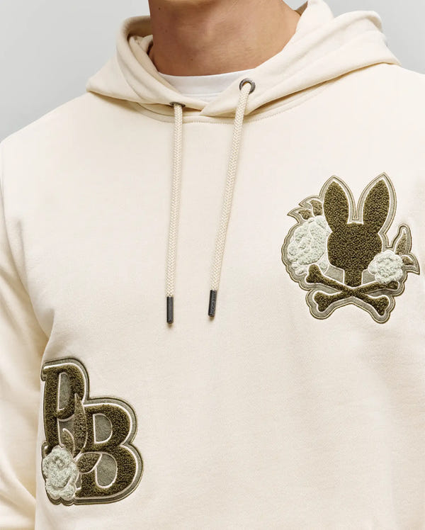 A person wears the Psycho Bunny MEN'S AUGUSTE HOODIE (MT0500043) in cream, featuring premium cotton, patch designs with stylized chenille "PB" floral detailing, and a bunny head with crossbones and flowers on the chest. Upper torso shown only.