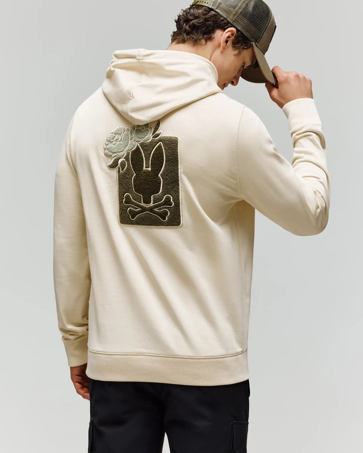 A man wearing the Psycho Bunny MENS AUGUSTE HOODIE (MT0500043) in beige and a green cap faces away, displaying a large black bunny, crossbones, and green rose chenille patch on the back.