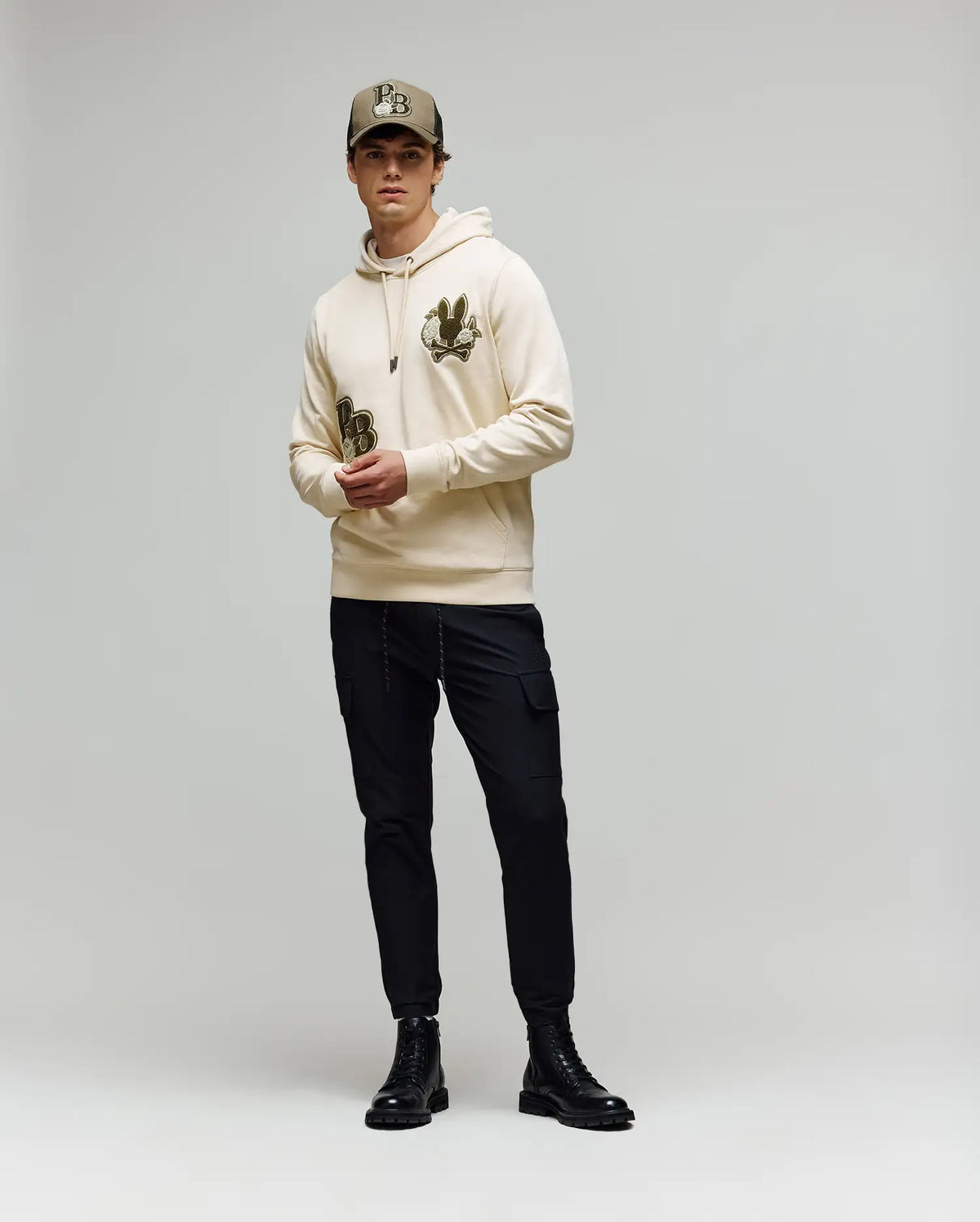 A man wearing the Psycho Bunny MENS AUGUSTE HOODIE - MT0500043, black cargo pants, black boots, and a beige baseball cap stands against a plain light gray background.