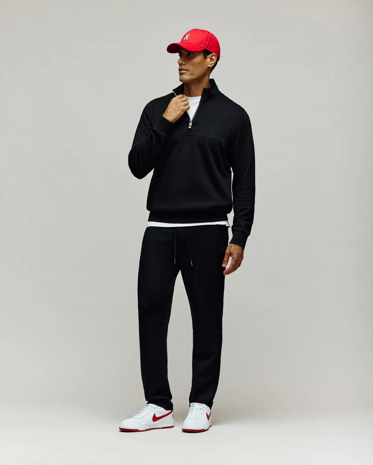 Mens Zeke 1/4 Mock Zip Pullover, a black classic fit French terry pullover featuring a silver 1/4 zip, mock neck with white lining, and a small embroidered black Psycho Bunny skull logo on the left chest, paired with black joggers and a red cap.