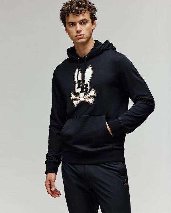 A young man with curly hair wears the Psycho Bunny MENS WADE HOODIE - MT0500112, featuring an embroidered Bunny skull and crossbones logo, paired with black pants, standing against a plain light background with one hand in his pocket.