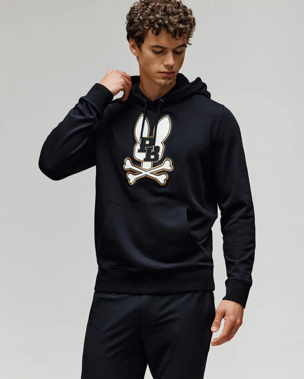 A young man with curly hair wears the Psycho Bunny MENS WADE HOODIE (MT0500112), a black classic athletic hoodie featuring an embroidered Bunny head and crossbones patch, as he looks down and touches its premium cotton blend collar.
