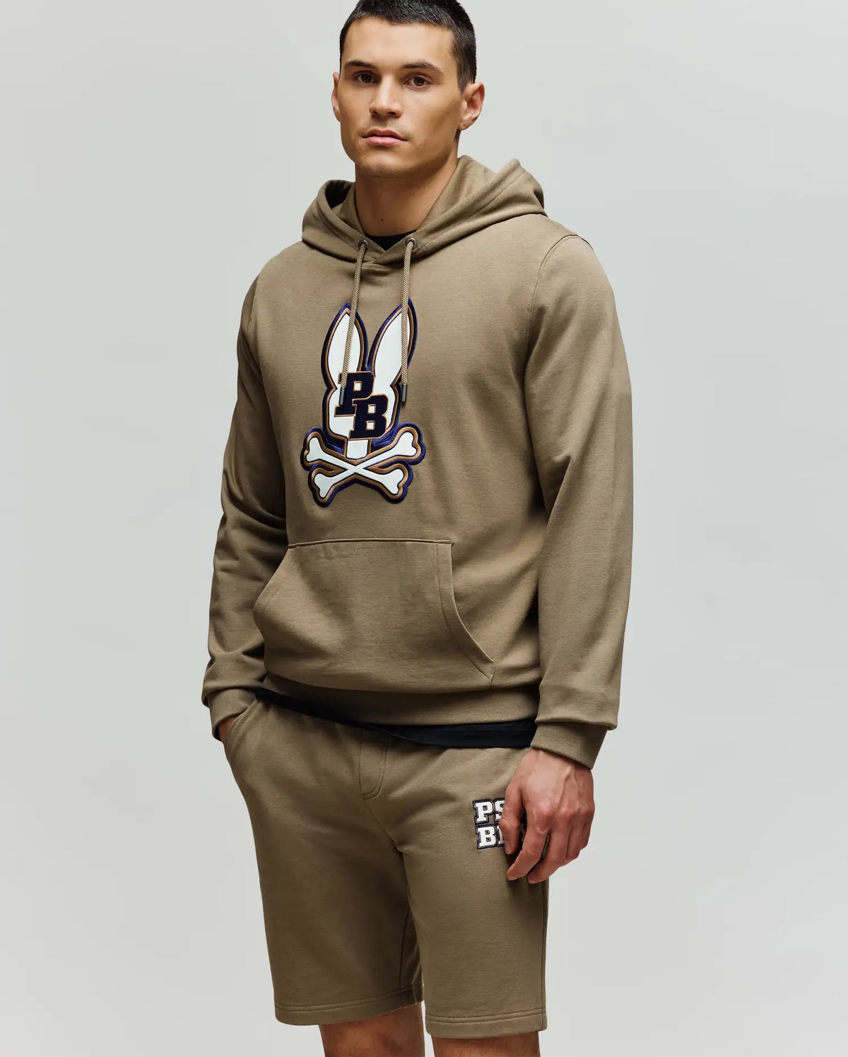 A man in a Psycho Bunny MENS WADE HOODIE (MT0500112) in khaki with an embroidered Bunny patch and matching khaki shorts featuring a logo on the left leg stands against a plain light background.