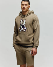 A man in a Psycho Bunny MENS WADE HOODIE (MT0500112) in khaki with an embroidered Bunny patch and matching khaki shorts featuring a logo on the left leg stands against a plain light background.
