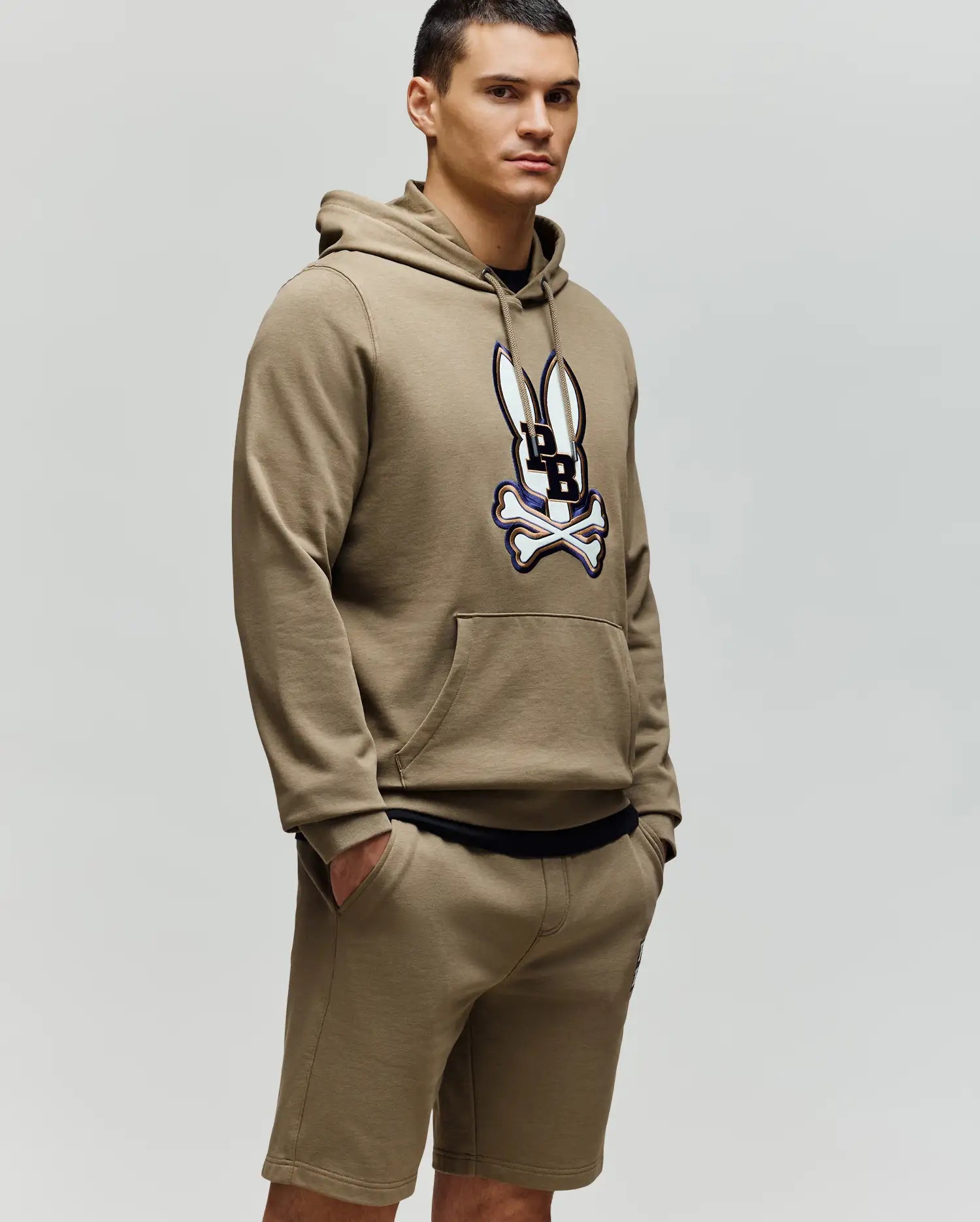 A man stands against a light grey background wearing the Psycho Bunny MENS WADE HOODIE (MT0500112), featuring an embroidered Bunny patch and crossbones on the front, paired with matching tan shorts. His hands are in his pockets as he looks to the side.