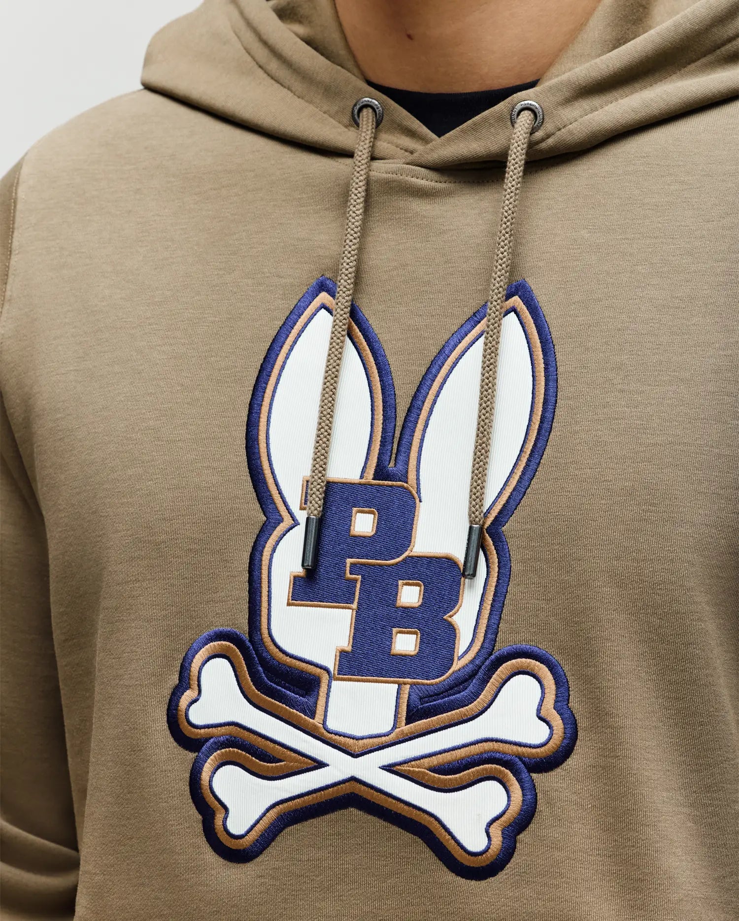 A man wears the Psycho Bunny MENS WADE HOODIE - MT0500112, a tan athletic hoodie made from a premium cotton blend, featuring an embroidered Bunny patch with ears, crossbones, and "PB" at the center.
