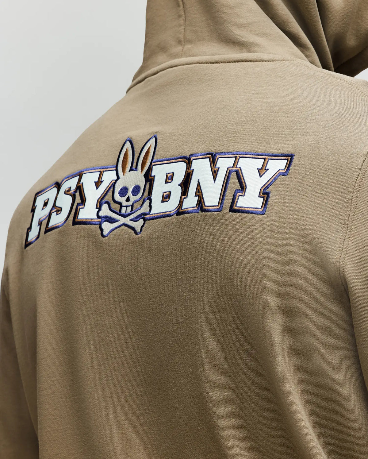 A person wearing the Psycho Bunny MENS WADE HOODIE - MT0500112 in tan, featuring "PSYBNY" boldly on the back and a skull-and-crossbones with bunny ears, made from a premium cotton blend.