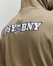 A person wearing the Psycho Bunny MENS WADE HOODIE - MT0500112 in tan, featuring "PSYBNY" boldly on the back and a skull-and-crossbones with bunny ears, made from a premium cotton blend.