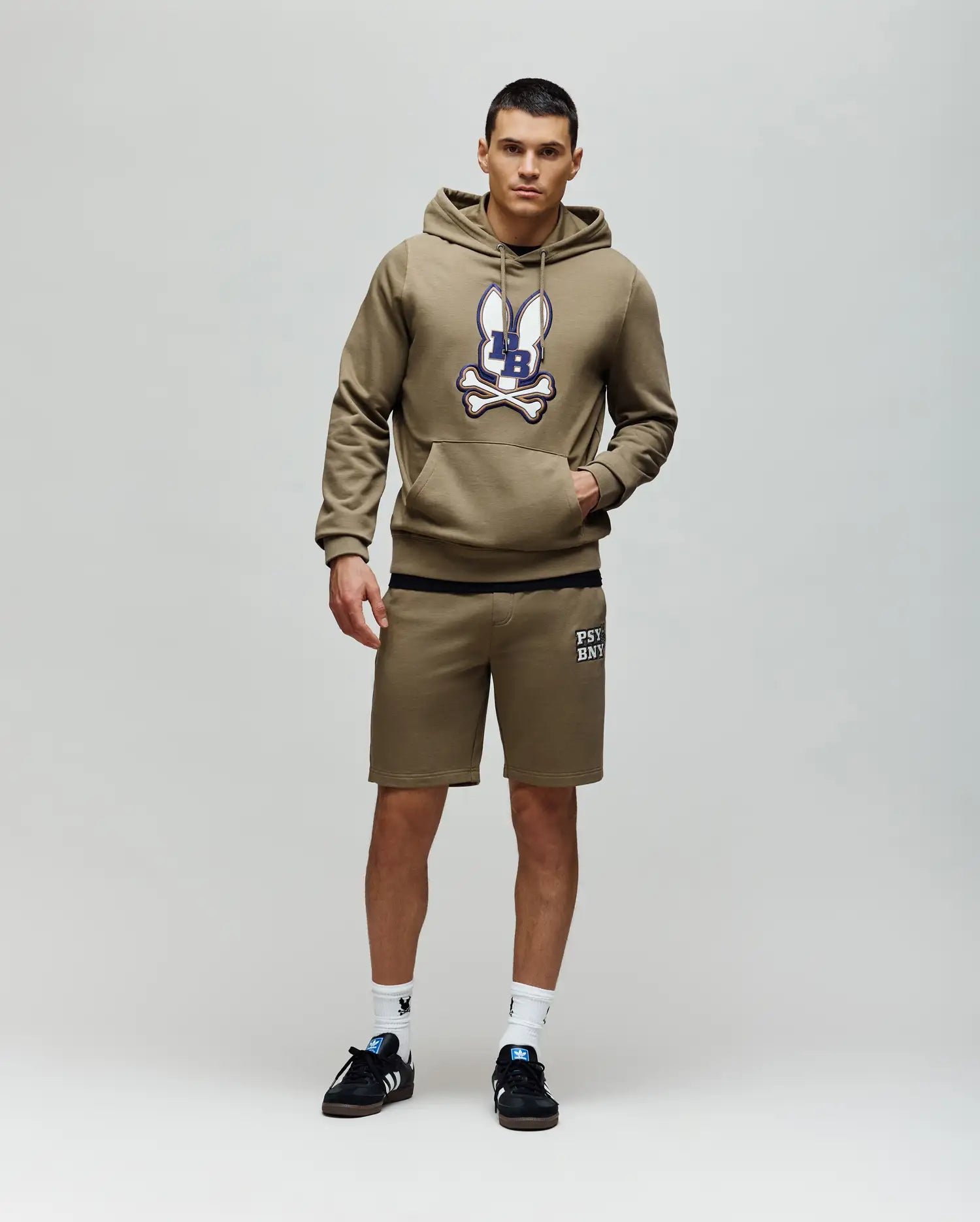 A man stands against a plain background wearing the Psycho Bunny MENS WADE HOODIE - MT0500112, featuring an embroidered Bunny patch, paired with matching khaki shorts, black sneakers, and white socks.