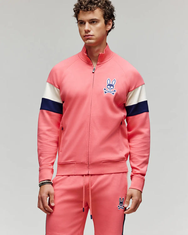 A man with curly hair models Psycho Bunny’s MENS DAWSON TRACK JACKET - MT0500113 and matching shorts in pink with navy and white stripes plus the bunny skull logo, standing against a plain light gray background.