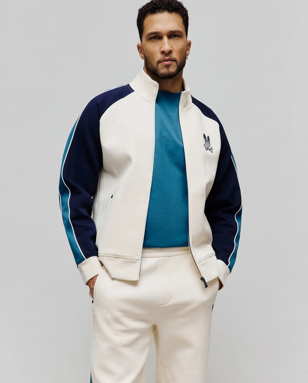 Mens Caspian Track Bomber in cream with navy blue sleeves, teal stripes, and white piping, featuring a classic fit, smooth fabric, and an embroidered Psycho Bunny skull logo on the left chest.