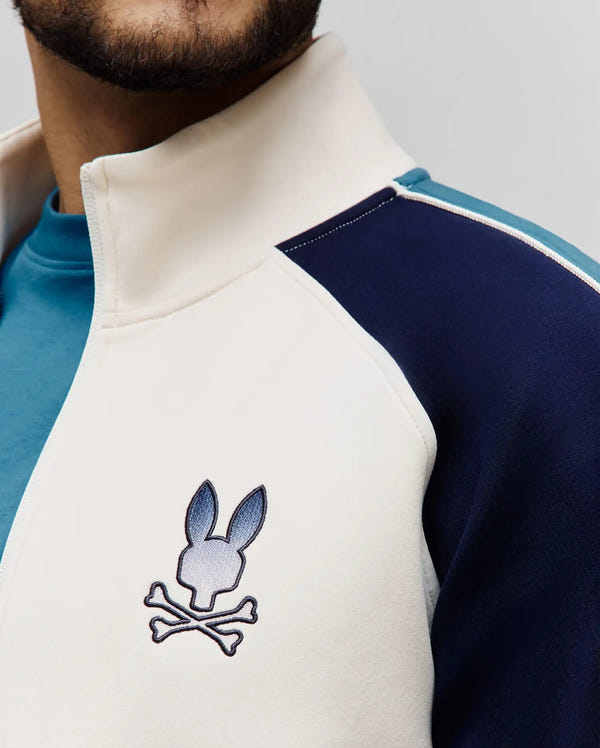 Mens Caspian Track Bomber in cream, navy, and light blue color-block design with white piping, featuring a smooth fabric and an embroidered blue gradient Psycho Bunny skull logo on the left chest.