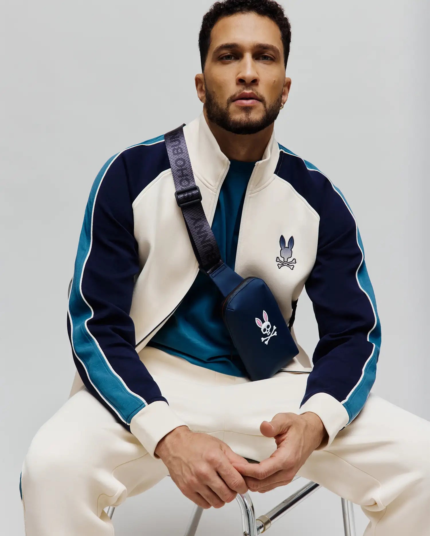 Mens Caspian Track Bomber in cream, navy, and teal color-block with white piping, featuring a classic fit and a small embroidered Psycho Bunny skull logo on the left chest.
