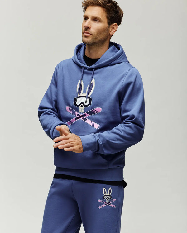 Mens Indigo Ski Bunny Hoodie. Classic fit indigo hoodie with a large embroidered white, black, and pink ski bunny skull logo on the chest, featuring ski goggles and crossed skis. Smooth jersey fabric, drawstring hood, ribbed cuffs.