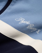 Men's Jacques Pique Track Jacket in light blue and navy pique fabric with a white stripe, featuring an embroidered white Psycho Bunny script logo and small bunny skull logo on the chest. (7)