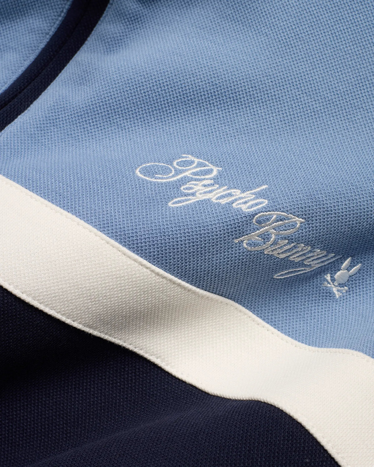 Men's Jacques Pique Track Jacket in light blue and navy pique fabric with a white stripe, featuring an embroidered white Psycho Bunny script logo and small bunny skull logo on the chest. (7)