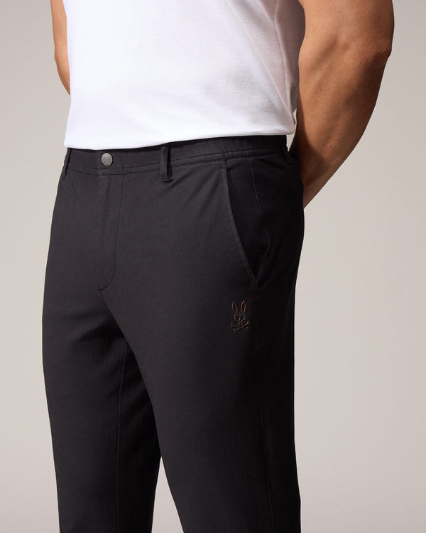 A close-up of a man in a light background wearing dark, tailored Psycho Bunny MENS MADISON COMMUTER PANTS with embroidered Bunny logos, focusing on the fit and muscular build of his lower body and biceps.