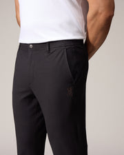 A close-up of a man in a light background wearing dark, tailored Psycho Bunny MENS MADISON COMMUTER PANTS with embroidered Bunny logos, focusing on the fit and muscular build of his lower body and biceps.