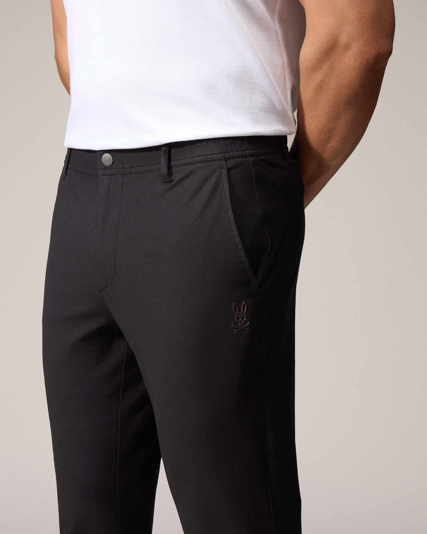 A close-up of a man in a light background wearing dark, tailored Psycho Bunny MENS MADISON COMMUTER PANTS with embroidered Bunny logos, focusing on the fit and muscular build of his lower body and biceps.
