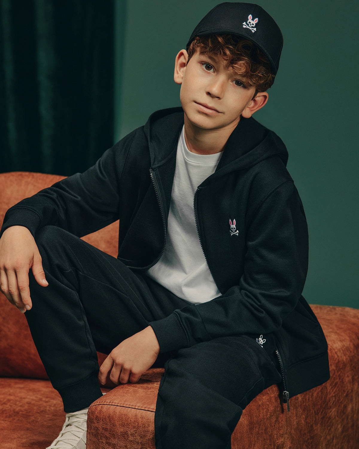 A young boy with curly hair sits casually on an orange couch, dressed in cozy fleece from head to toe. He sports a black Psycho Bunny cap with a rabbit logo, a black hoodie, KIDS LEVY ESSENTIAL FLEECE SWEATPANT - B0P721D200, and a white t-shirt. The dark green wall behind him adds depth to the scene.