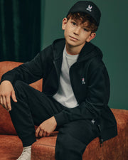 A young boy with curly hair sits casually on an orange couch, dressed in cozy fleece from head to toe. He sports a black Psycho Bunny cap with a rabbit logo, a black hoodie, KIDS LEVY ESSENTIAL FLEECE SWEATPANT - B0P721D200, and a white t-shirt. The dark green wall behind him adds depth to the scene.