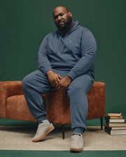 A man in a blue Psycho Bunny MENS BIG AND TALL LEVY ESSENTIAL FLEECE HOODIE (B9H859D200) and matching sweatpants sits on a brown chair, looking at the camera. He wears gray sneakers. The green background has stacks of books beside him.