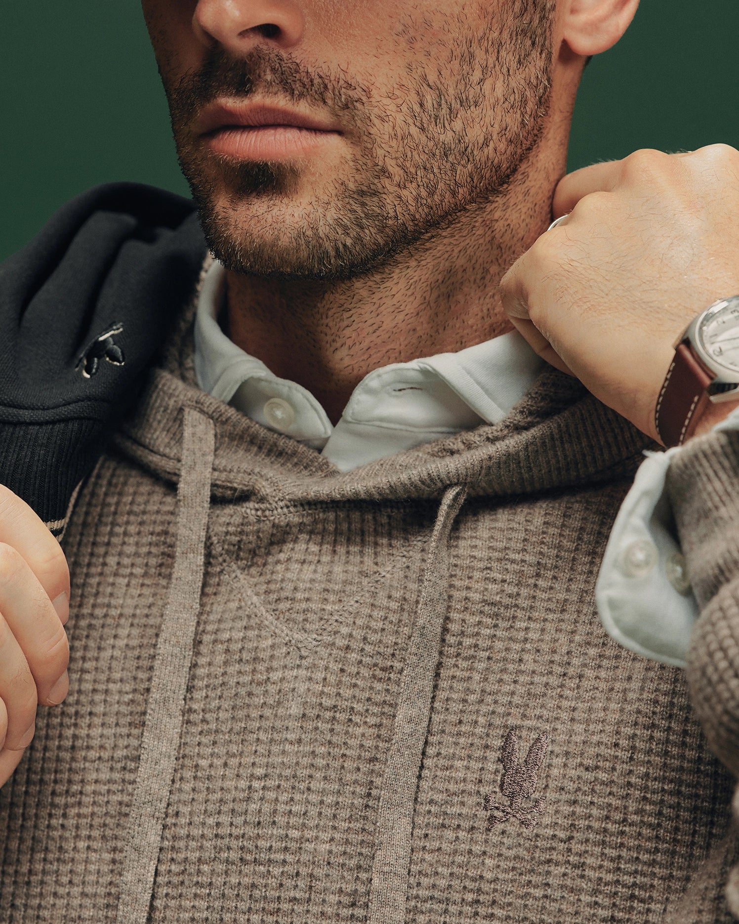 A man in a Psycho Bunny MENS LEVAN WAFFLE HOODED SWEATER adjusts the hoodie over a white collared shirt. With a neatly trimmed beard, he shows off a watch on one hand against a green background.