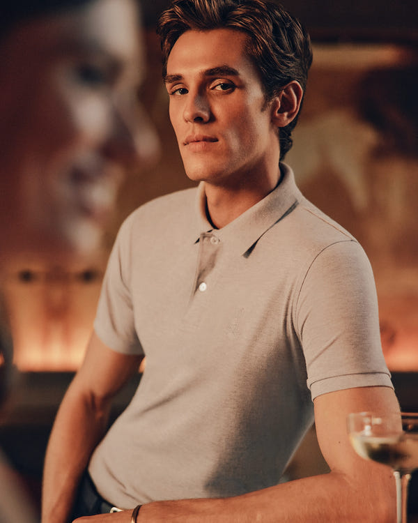 A person in a Psycho Bunny MENS JAMES PREMIUM PIQUE POLO SHIRT leans against a bar counter with confidence. A blurred figure is in the foreground, and there's a glass of white wine on the counter. The background features a warm, soft focus.