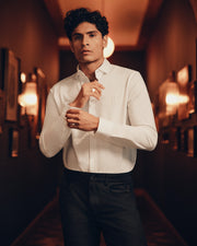 In a dim hallway adorned with framed pictures, a person with short, curly hair adjusts their cuff while wearing Psycho Bunny's crisp white MENS ARTHUR JERSEY SHIRT - B6C748D200, crafted from stretch Pima cotton, paired with black pants. A round light fixture softly glows in the background.