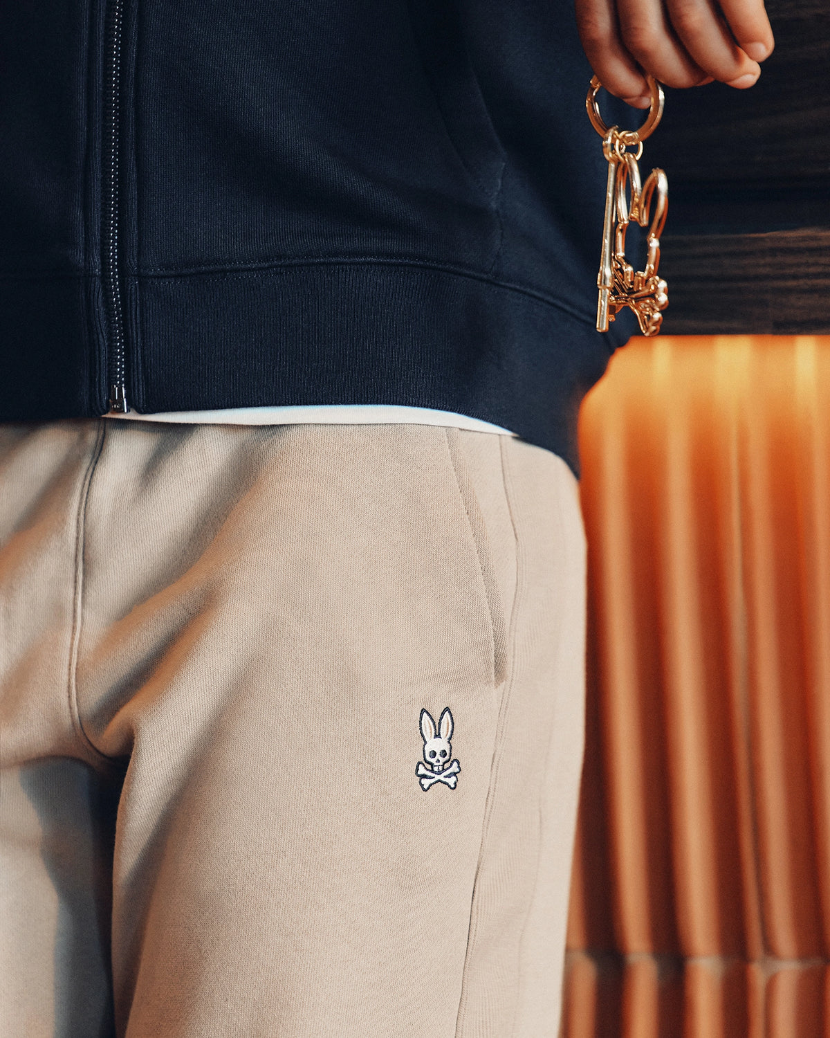 Close-up of a person holding gold keys adorned with a small bear charm. They're wearing the KIDS LEVY ESSENTIAL FLEECE SWEATPANT by Psycho Bunny and a beige garment featuring their stitched cartoon bunny logo with crossed bones below it. A dark jacket partially obscures the background, enhancing the ensemble's relaxed vibe.