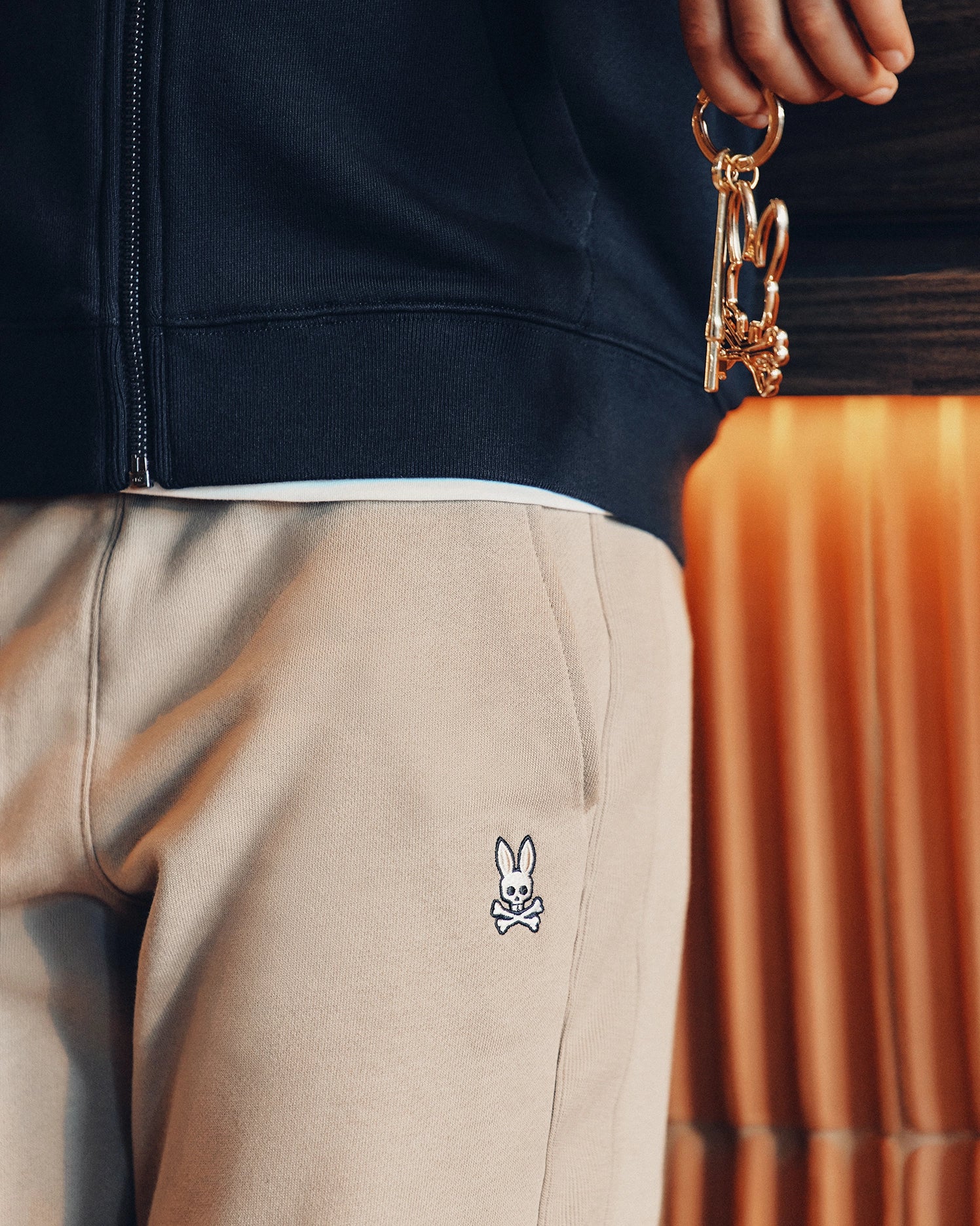 Close-up of a person holding gold keys adorned with a small bear charm. They're wearing the KIDS LEVY ESSENTIAL FLEECE SWEATPANT by Psycho Bunny and a beige garment featuring their stitched cartoon bunny logo with crossed bones below it. A dark jacket partially obscures the background, enhancing the ensemble's relaxed vibe.